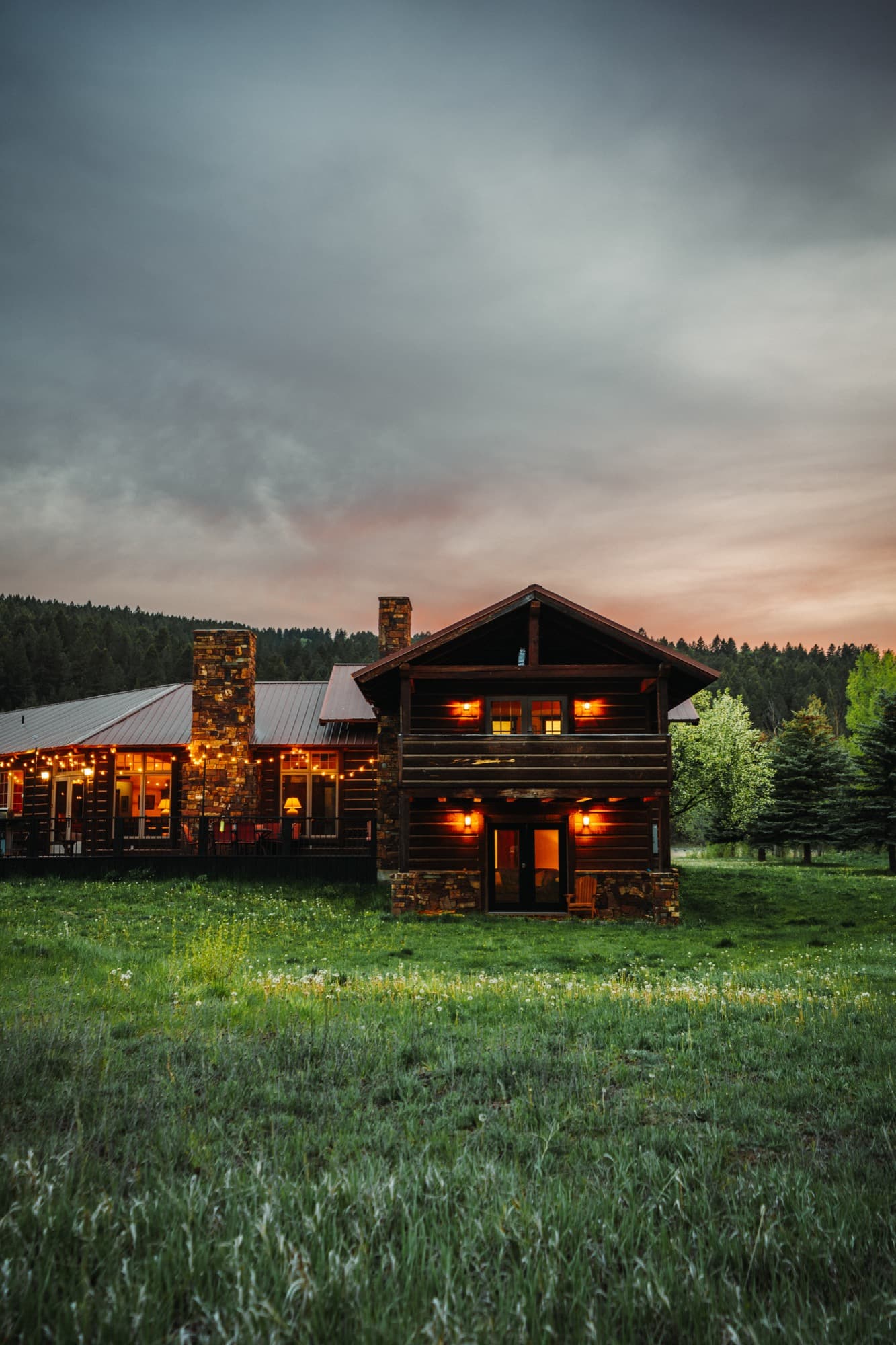 Two-story log cabin at dusk with warm lit windows under a dramatic sky